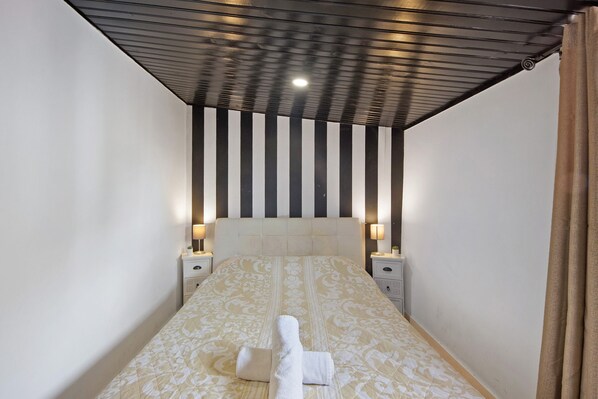 A narrow hotel room with a bed, bedside lamps, a headboard with vertical stripes, and a curtain on the right.