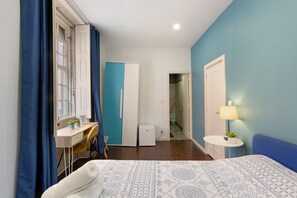 A bedroom with a bed, a desk, a chair, a small table with a lamp, and a wardrobe.
