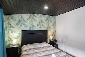 Een slaapkamer met een bed, een nachtkastje, een tafeltje met een lamp, een ventilator aan de muur en een plafondventilator.
