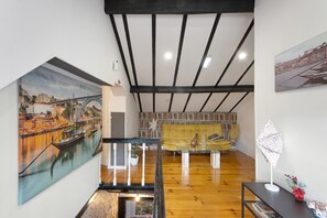 A room with a wooden floor, a staircase, a sofa, a table, and a painting of a bridge.