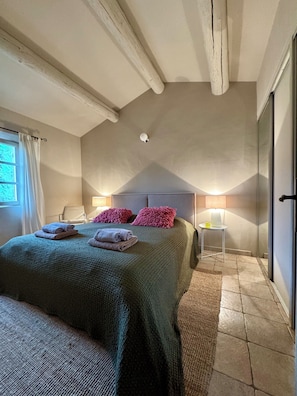 Un dormitorio con cama, almohadas, mesita de noche, lámpara y ventana con cortinas.