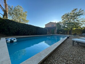 Ein rechteckiger Swimmingpool mit einem Kiesstreifen am Rand, umgeben von einer Hecke und Bäumen.