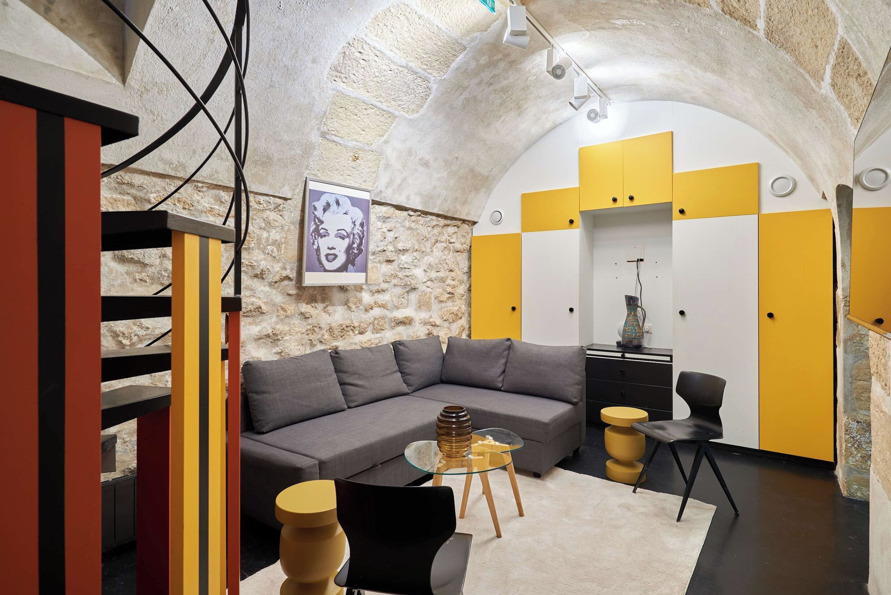 Un salon moderne avec un mur en pierre, un canapé gris, des meubles jaunes et blancs, et une table basse en verre.