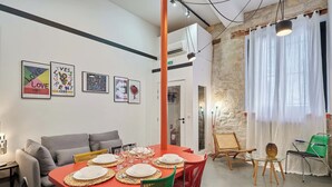 Un comedor con una mesa roja dispuesta para cuatro, rodeado de sillas coloridas, un sofá y una pared de piedra.