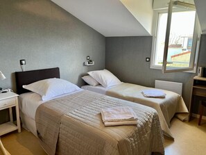 Habitación de hotel con dos camas, una mesita de noche, una lámpara, una estantería con una planta pequeña y una ventana con vistas al exterior.