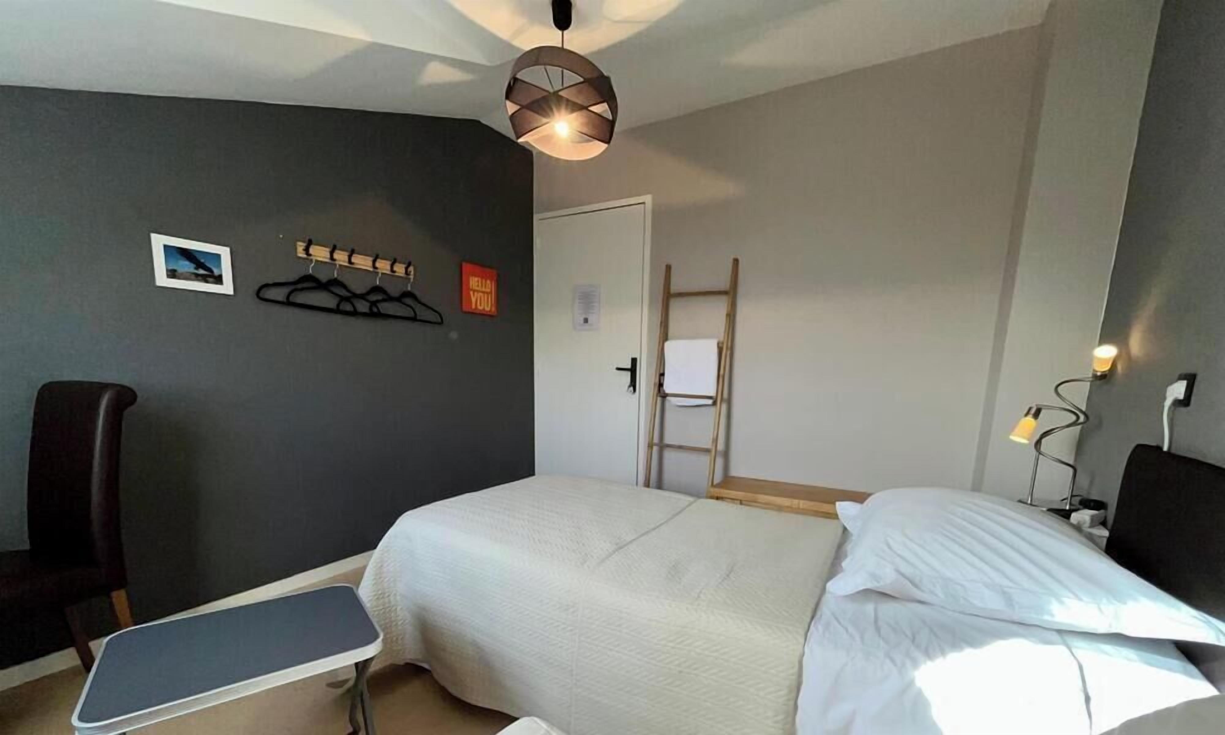Une chambre d’hôtel avec un lit, une chaise, un porte-manteau, une échelle et un bureau avec une lampe.