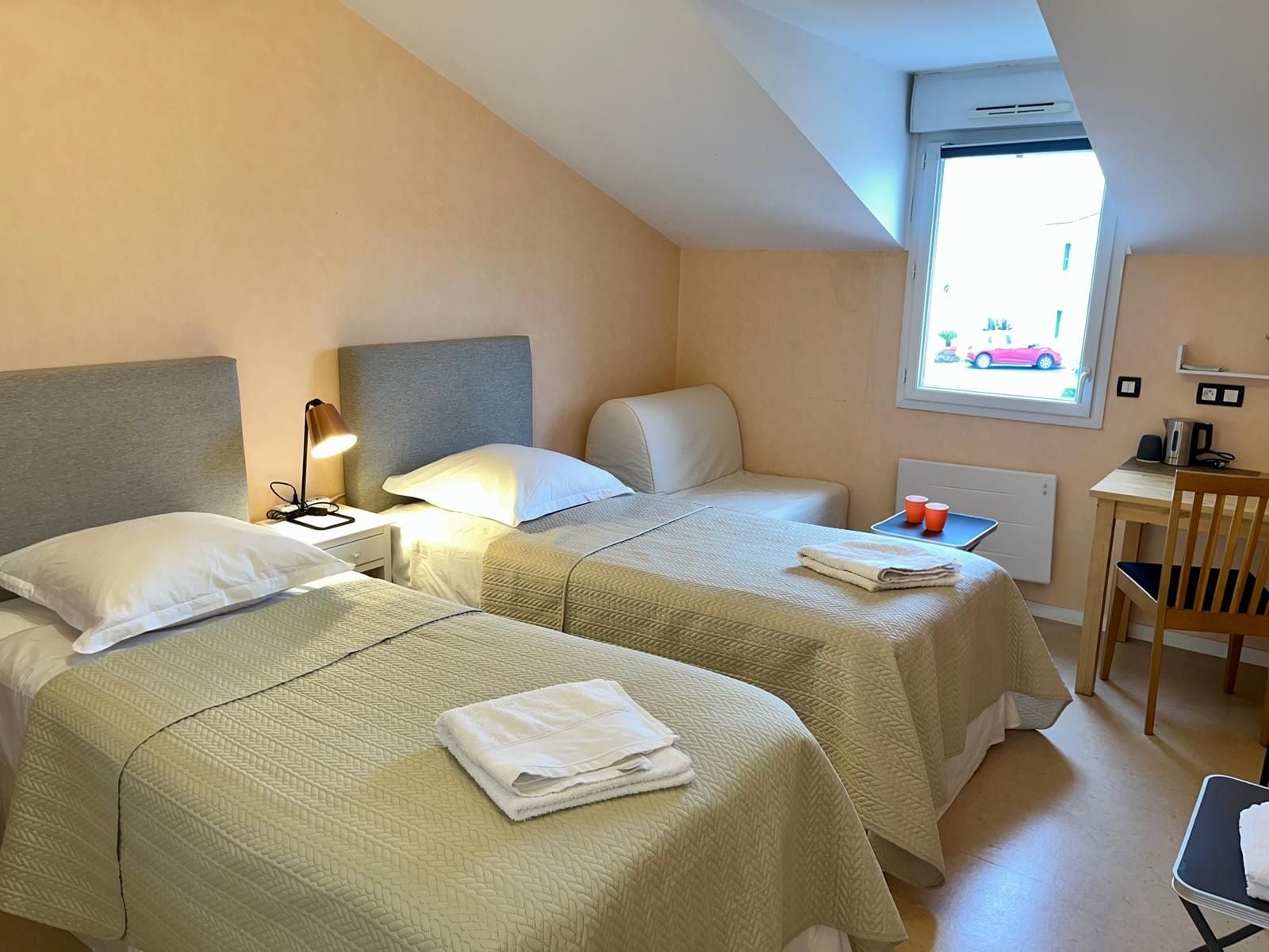 Een hotelkamer met twee bedden, een bureau, een stoel en een raam met uitzicht op een auto.