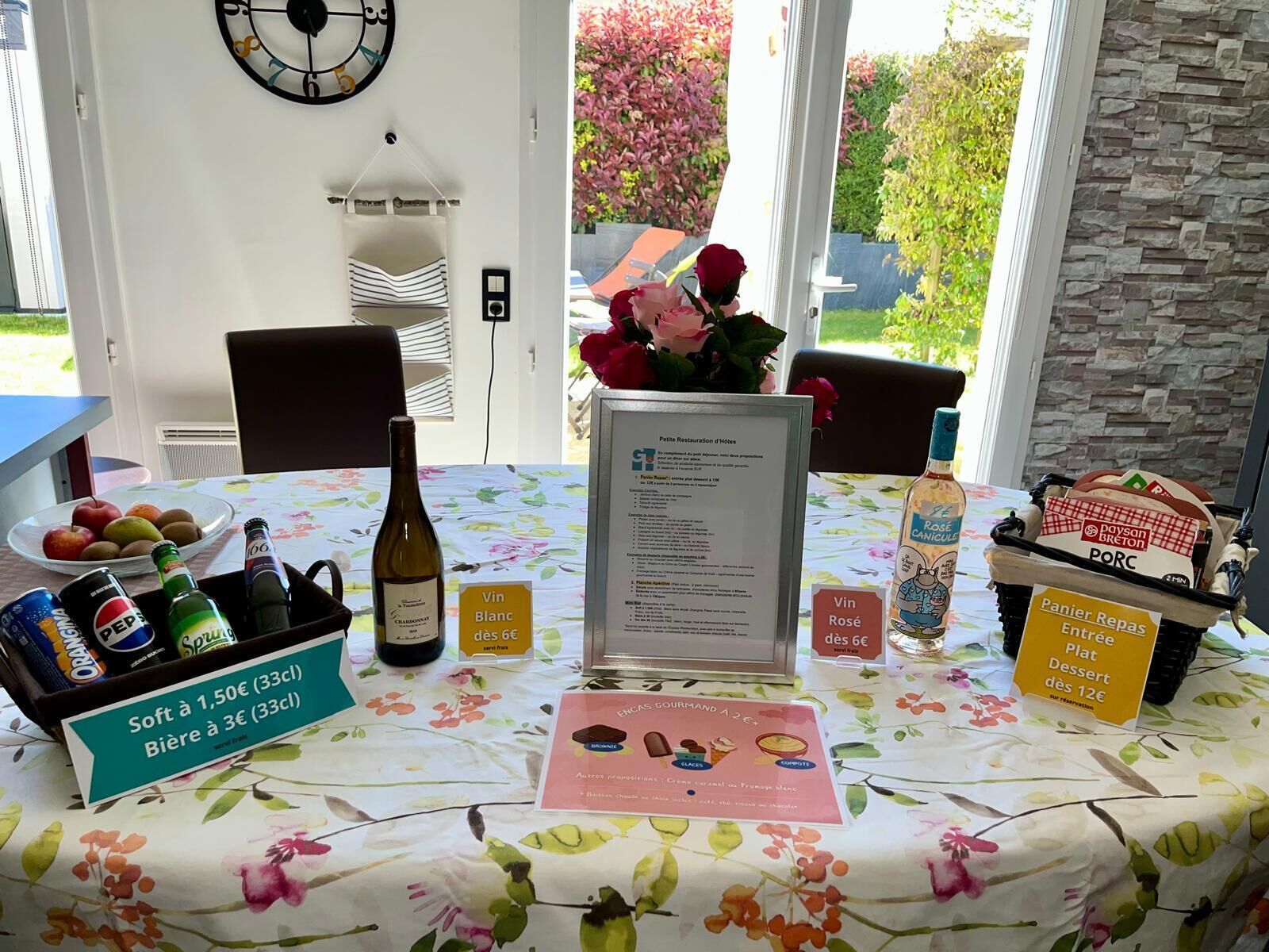 Een tafel met een tafelkleed met bloemen, een menukaart, een fles wijn, een mand met snacks en een vaas met bloemen.