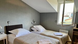 Una habitación con dos camas, un escritorio, una silla, una ventana y un pequeño estante con un cuadro.