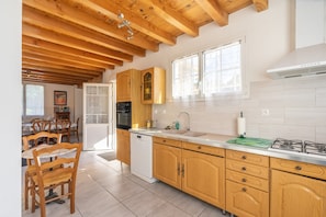 A kitchen with wooden cabinets, a dishwasher, a stove, and a dining area with chairs and a table. - Bright Apartment 300m from the Beach (La Tremblade)