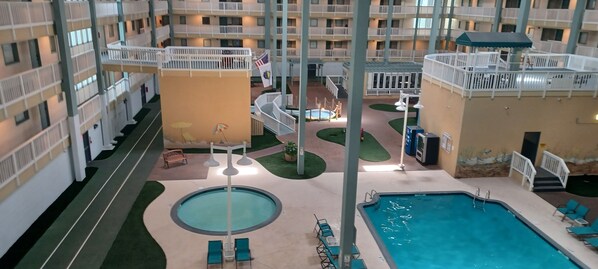 A hotel atrium with multiple balconies, a central pool area with lounge chairs, and a children's play area.
