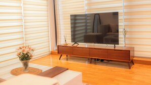 Ein modernes Wohnzimmer mit einem großen Flachbildfernseher an einer Holzkonsole, einer Vase mit Blumen auf einer gewebten Matte und einem Sofa im Vordergrund.