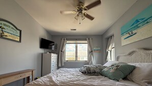 Un dormitorio con cama, ventilador de techo, televisor, cómoda y ventana con cortinas.