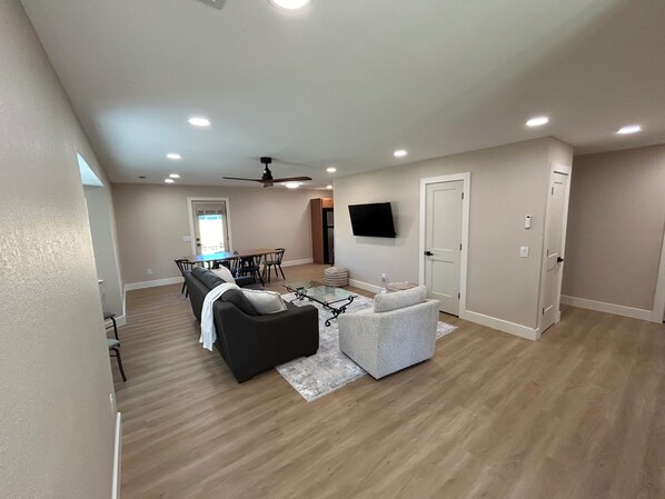 A spacious living room with a dark sofa, a light-colored armchair, and a glass coffee table. A dining area with a table and chairs is visible in the background.