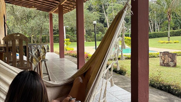 Una hamaca bajo una pérgola de madera con piso de baldosas, rodeada de follaje y una piscina.