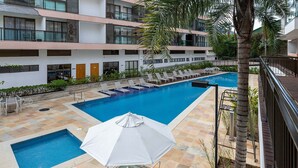 A pool area with multiple lounge chairs and umbrellas, surrounded by a multi-story building with balconies and windows.