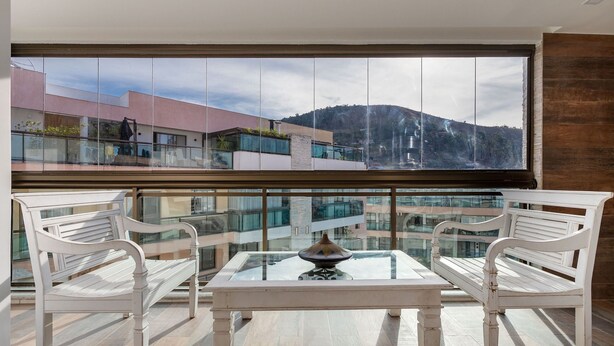 A balcony with a glass table and two white chairs, overlooking a modern building and a mountainous landscape.