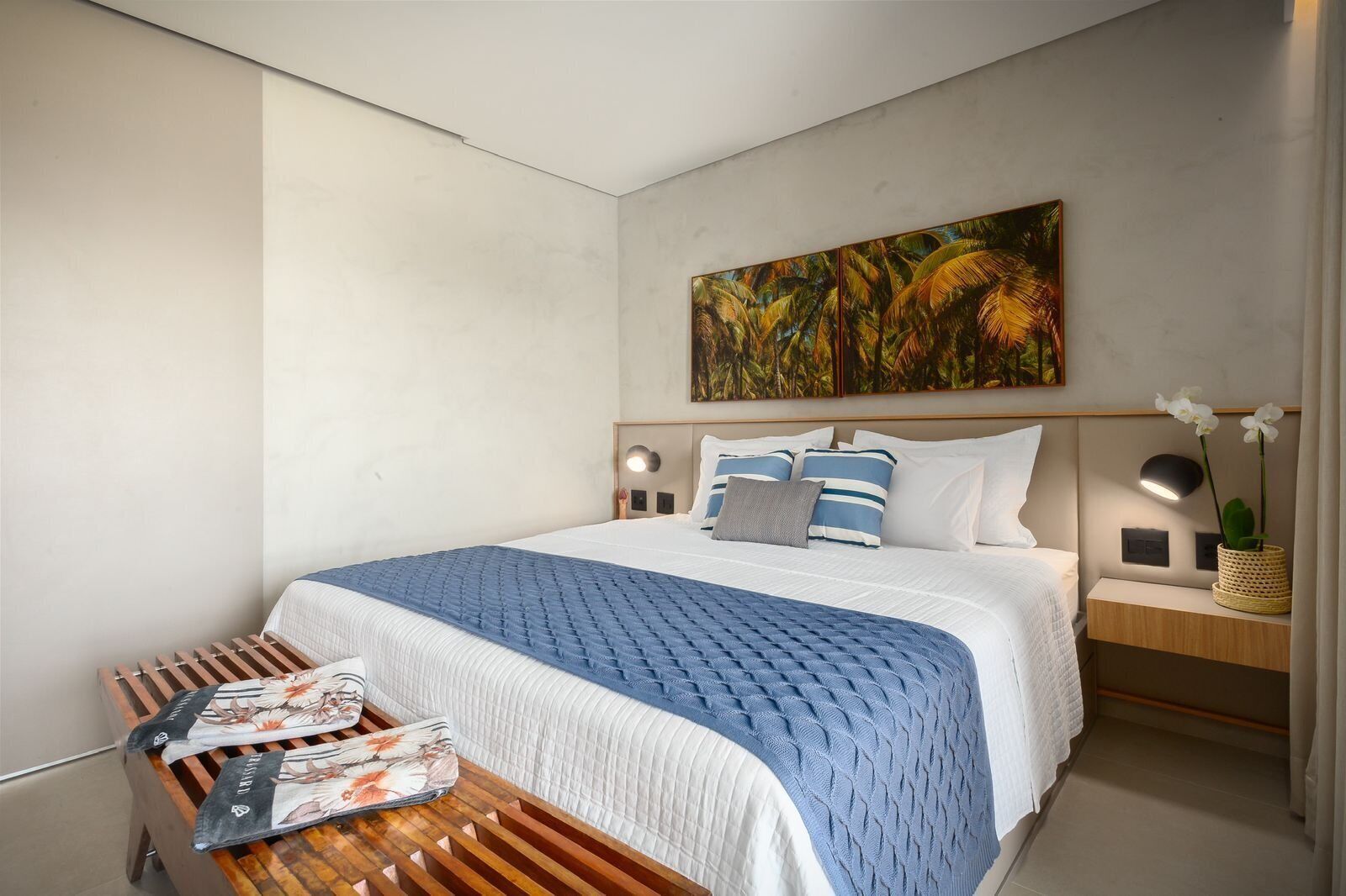 A modern hotel room with a large bed, a wooden side table, a nightstand with a lamp, and a painting above the bed.