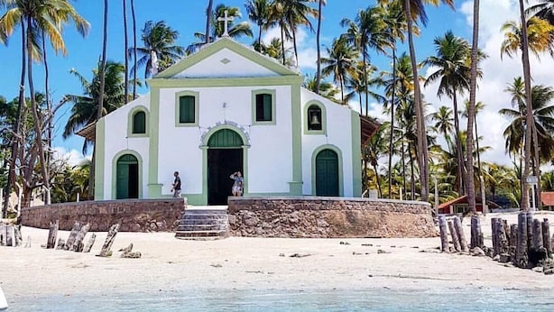 Una iglesia blanca con detalles verdes, rodeada de palmeras y una playa.