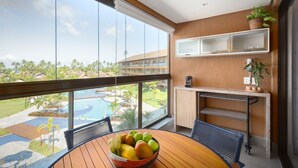 Un balcón con una mesa de madera, un cuenco de fruta y vistas a la piscina de un resort y palmeras.