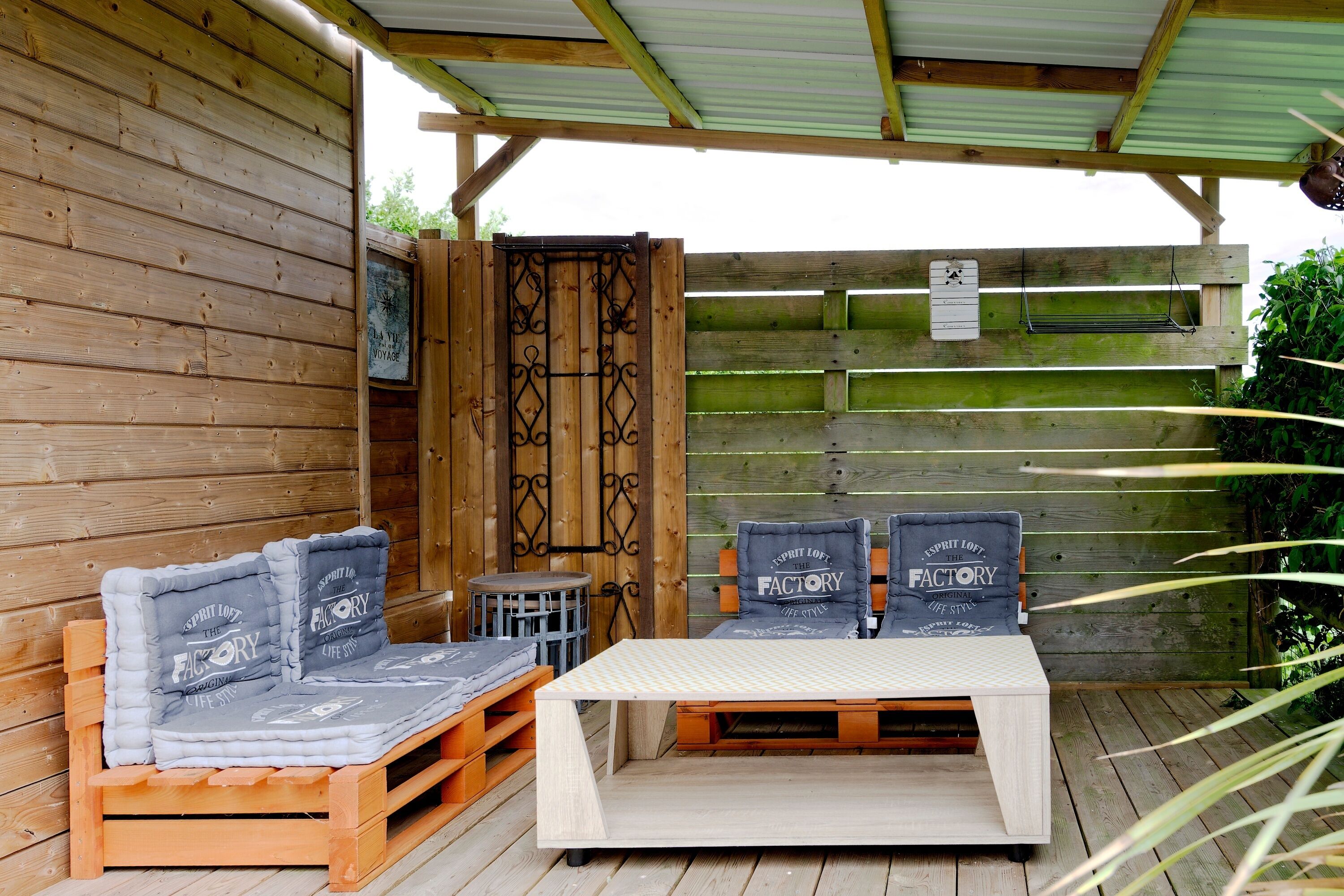 A wooden deck with a wooden bench and a small table, a covered area with a metal roof, and a wooden fence with a sign.