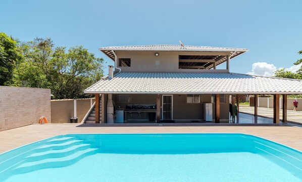 Une piscine avec un patio couvert, un mur en briques et un ciel d’un bleu limpide.