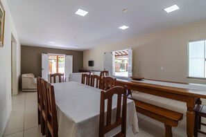 Un espace repas avec une longue table en bois, plusieurs chaises et un banc. On y trouve des luminaires au plafond, un téléviseur et une porte donnant sur l’extérieur.