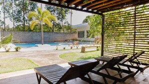 Un espacio exterior cubierto con sillones de madera, una piscina y vistas a una casa y árboles.