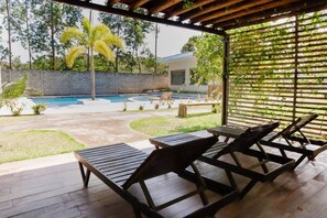A covered patio with wooden lounge chairs, a swimming pool, and a view of a house and greenery.
