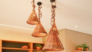 Una cocina moderna con armarios de madera, un refrigerador de acero inoxidable y una mesa de comedor con sillas. Hay luces colgantes de cobre y plantas en macetas sobre estantes.