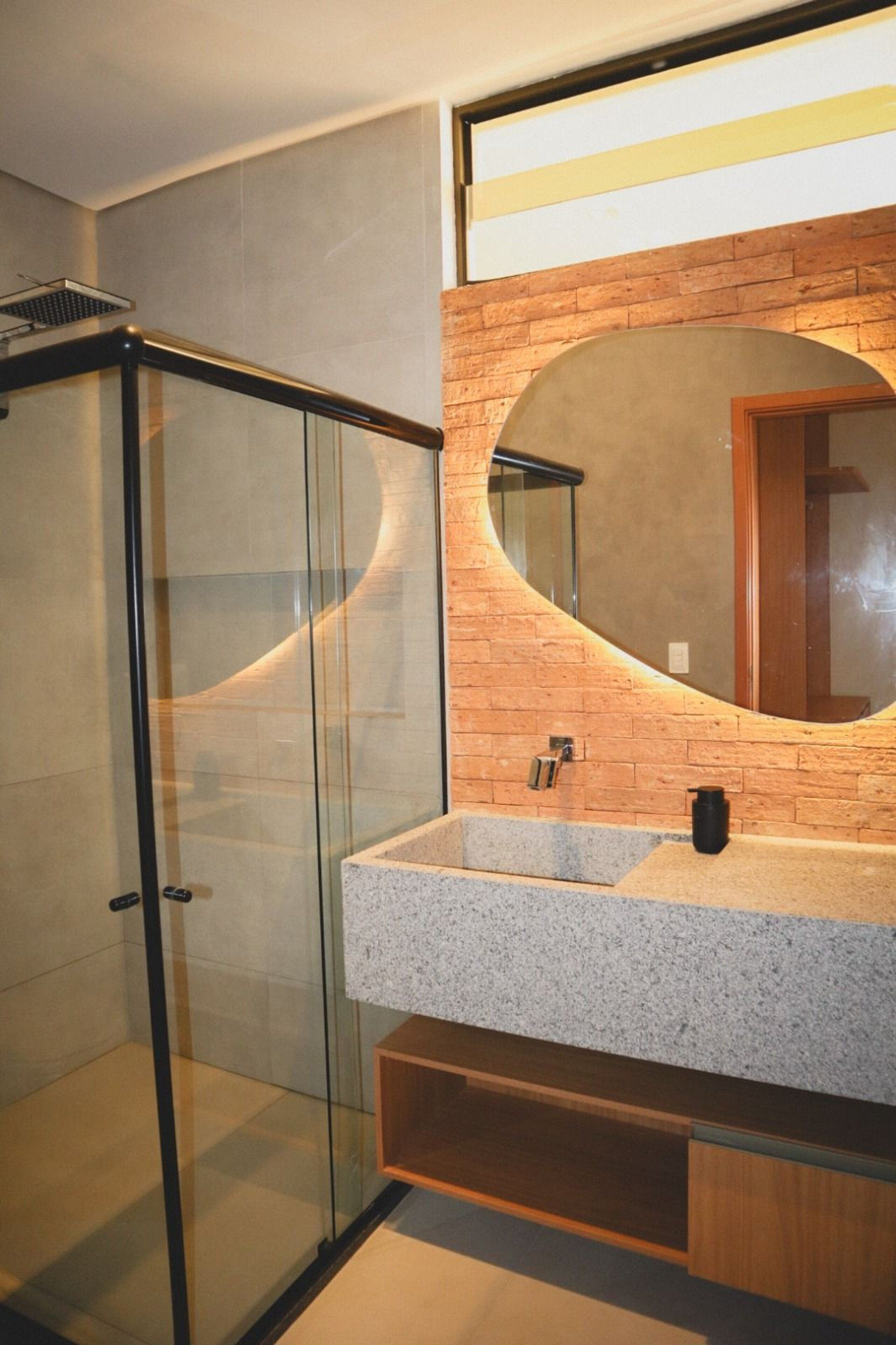 A modern bathroom with a glass shower enclosure, a granite countertop, and a large mirror with a brick wall backdrop.