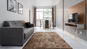 Un salon moderne avec un canapé gris, un tapis marron, un coin repas comprenant une table et des chaises, et un téléviseur fixé sur un panneau en bois.