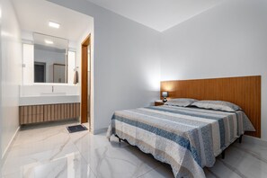 A modern bedroom with a large bed, a wooden headboard, a nightstand, a bedside lamp, and a bathroom with a mirror and sink.