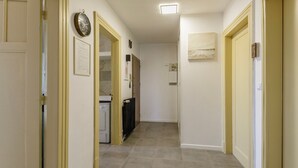 A narrow hallway with beige tiled flooring, cream walls, and yellow trim. There are doors on both sides, a radiator, and a clock on the wall.