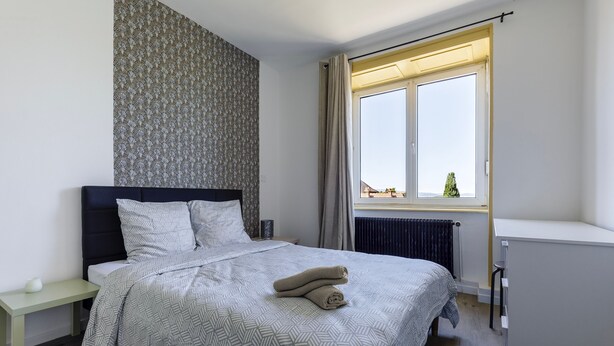 A bedroom with a bed, a nightstand, a radiator, a window with curtains, and a view of a tree outside.