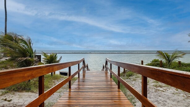 A wooden boardwalk leading to a beach with palm trees and a clear sky. - Flat beach front Itah Residence – Muro Alto/PE (Muro Alto)