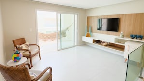A spacious living area with a large TV, wicker furniture, and a glass door leading to a balcony. - House with Private Pool in Maragogi – Praia de Antunes (Maragogi)