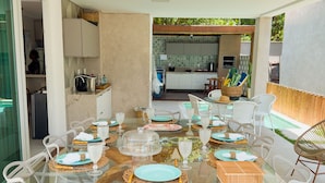 A glass-top dining table with white chairs, a kitchen in the background, and a view of the outdoors. - House with Private Pool in Maragogi – Praia de Antunes (Maragogi)