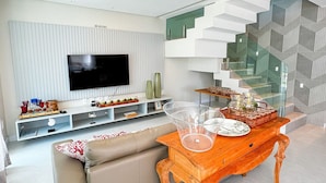 A modern living room with a flat-screen TV, a wooden coffee table, a beige sofa, and a staircase with a geometric design. - House with Private Pool in Maragogi – Praia de Antunes (Maragogi)