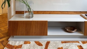 A modern living room with a flat-screen TV mounted on a white wall, a wooden console table with a glass vase containing a plant, and a patterned rug on the floor.