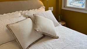 A bed with white bedding and decorative pillows, a bedside lamp, and a window with a view of buildings and hills.