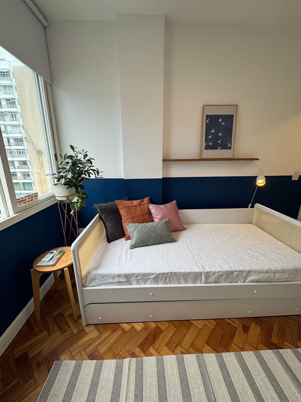 Un dormitorio con cama, una mesita de noche pequeña, una planta en maceta, un cuadro en la pared y una ventana con vista a edificios.