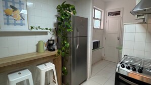 A kitchen with white tiled walls, a stainless steel refrigerator, a gas stove, and a microwave. There is a small stool and a potted plant on the counter.