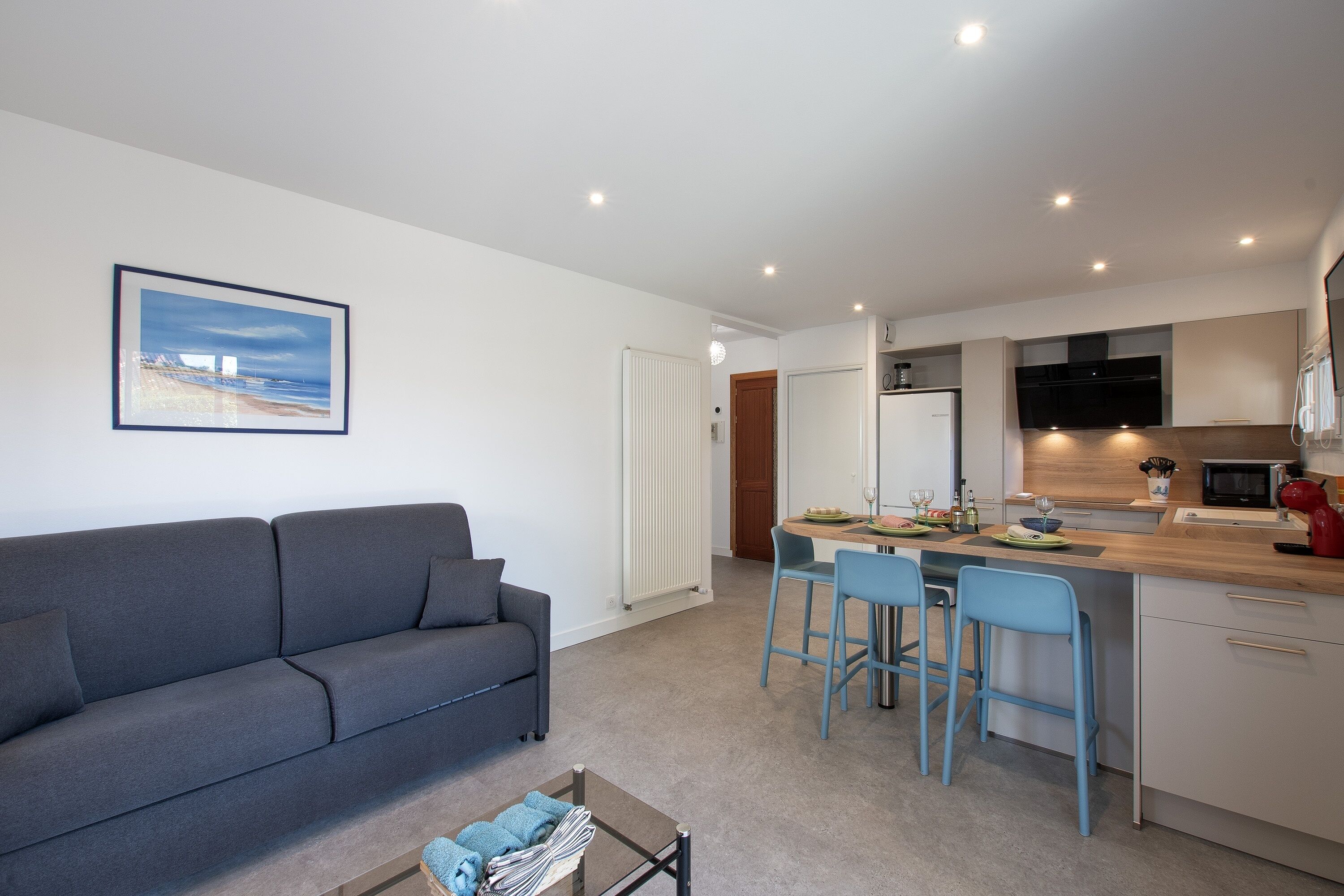 A modern apartment with a grey sofa, a dining area with blue chairs, and a kitchen with white cabinets.