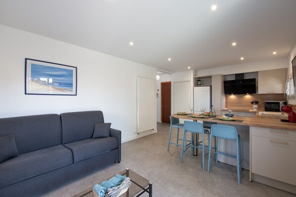 A modern apartment with a grey sofa, a dining area with blue chairs, and a kitchen with white cabinets.