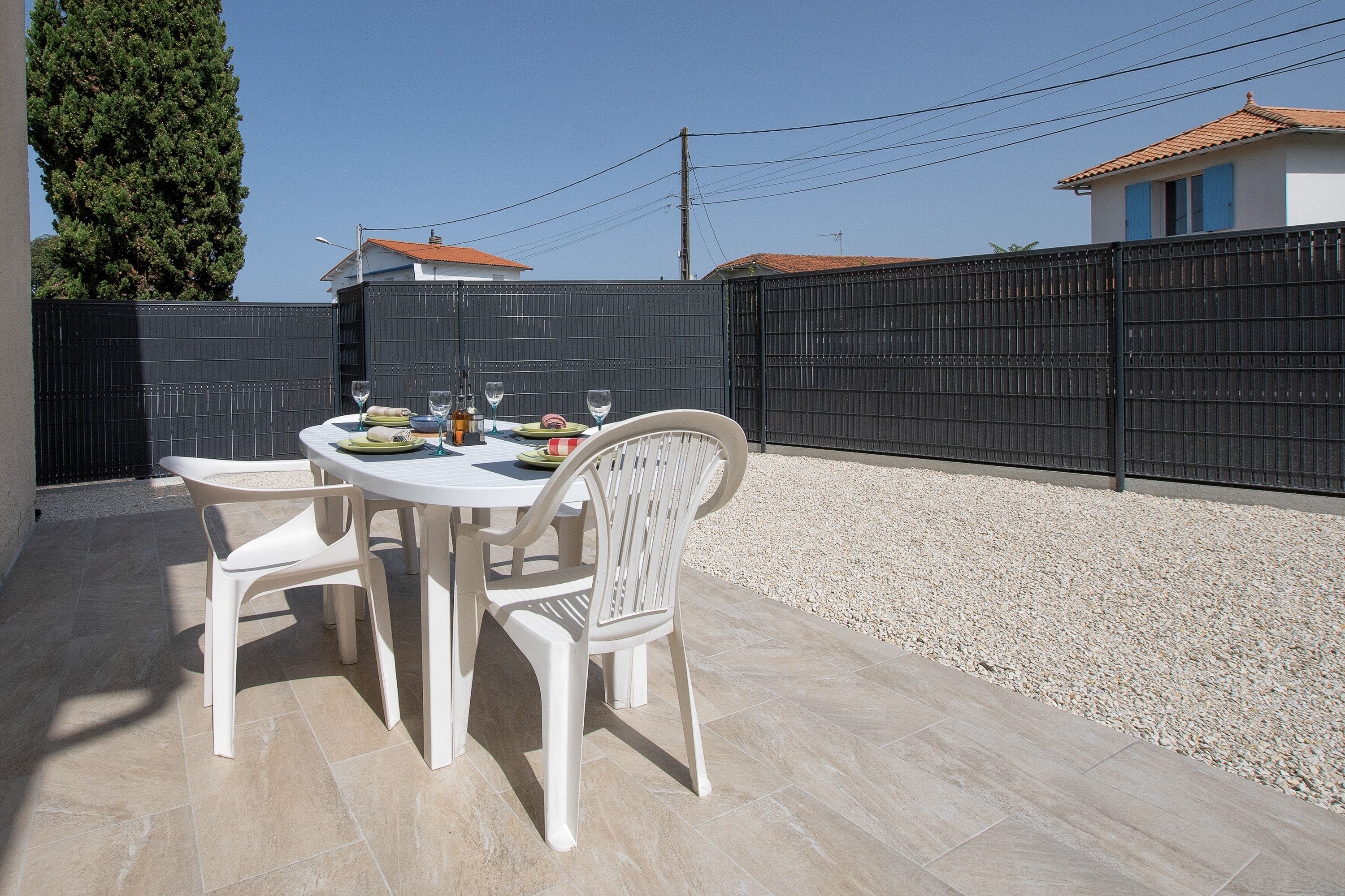 An outdoor patio with a round table and four white chairs, surrounded by a black fence and a gravel area.