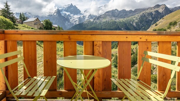 A wooden deck with a round table and two chairs, overlooking a mountainous landscape with snow-capped peaks and a village in the background.
