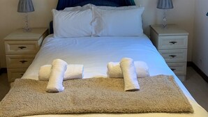 A neatly made bed with white linens, a beige throw blanket, and two towels folded at the foot of the bed. There are two bedside tables with lamps, and two framed pictures on the walls.