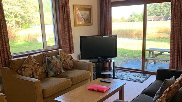 A living room with a sofa, armchairs, a coffee table, and a TV. There is a large window with curtains, a painting on the wall, and a view of a grassy area outside.