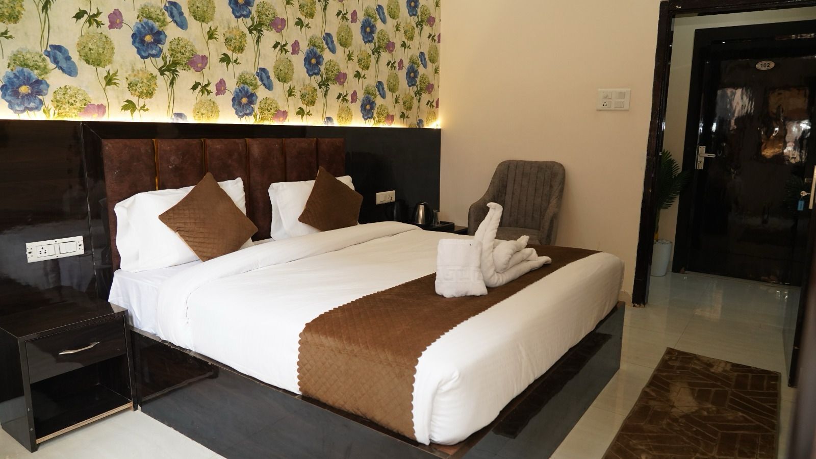 Una cama bien hecha con una manta marrón y una toalla doblada encima, una mesita de noche con una lámpara, y una silla en una habitación de hotel con papel tapiz floral.